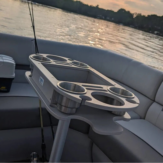A gray and stainless steel boat table with integrated cup holders and rod holders, shown on a boat with water and trees in the background at sunset.