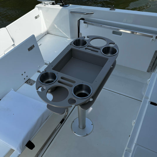 Gray adjustable boat table with integrated cup holders and a central storage compartment, mounted on a pedestal in a white boat cabin.