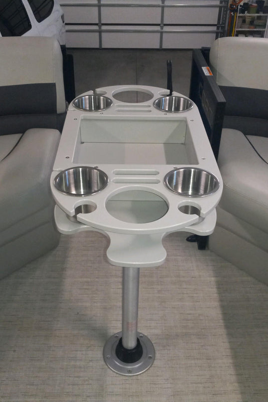 A white and silver center pedestal table with integrated cup holders and a glass top, positioned between two cream and dark gray upholstered boat seating sections.